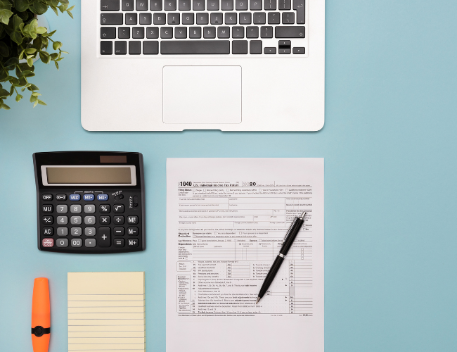 A laptop, a calculator, notepad, and a orange highlighter on a light blue table